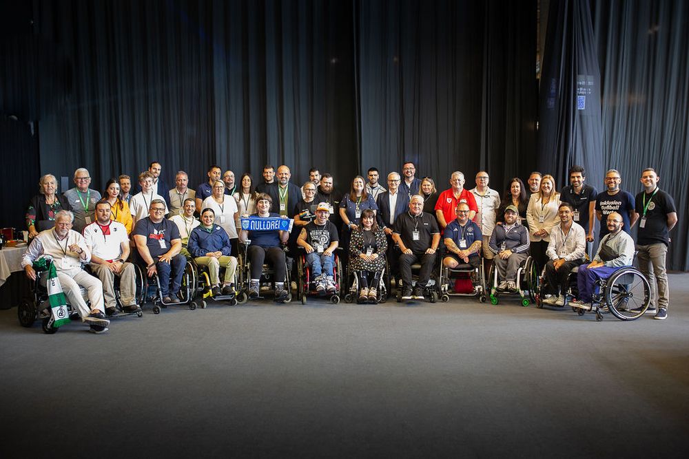 A large group photo of attendees from the Disability and Inclusion Fan Network event in Malmö. The diverse group includes many individuals in wheelchairs, standing together and smiling in front of a dark curtain backdrop. Several people wear football-related shirts.