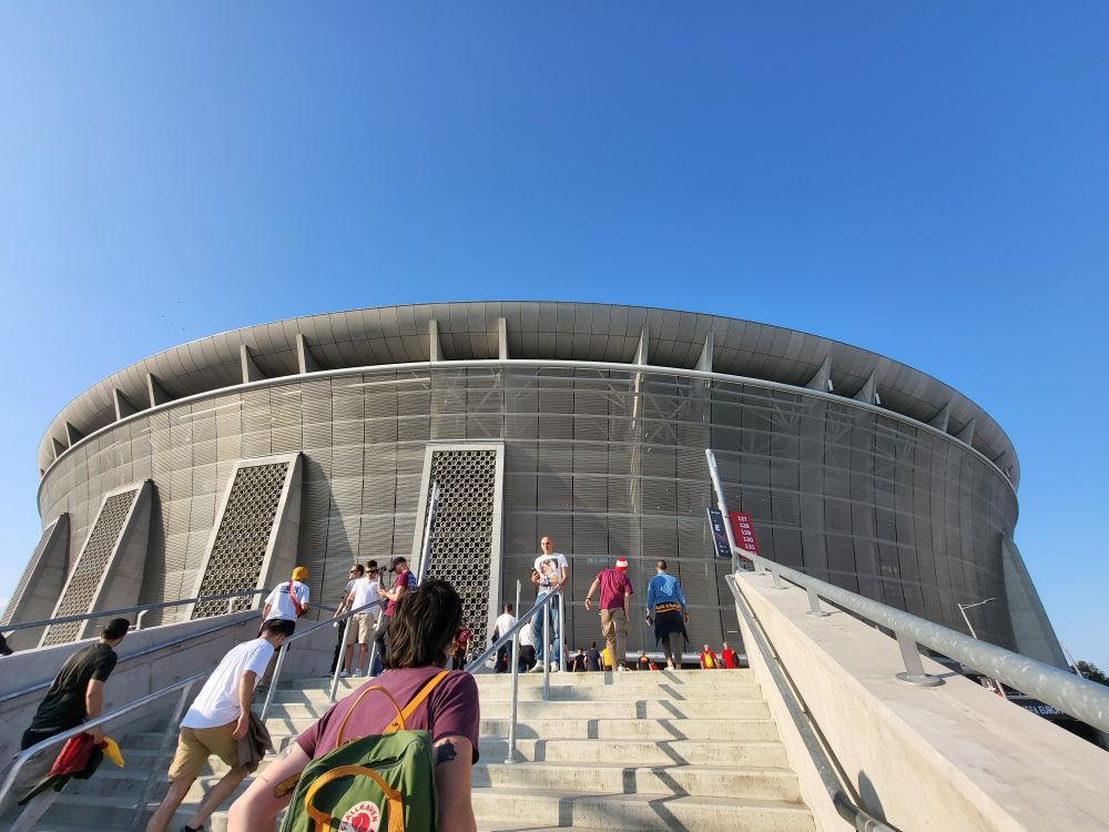 Fans walk up the steps towards a modern circular football stadium under a clear blue sky, creating a sense of anticipation ahead of the match.