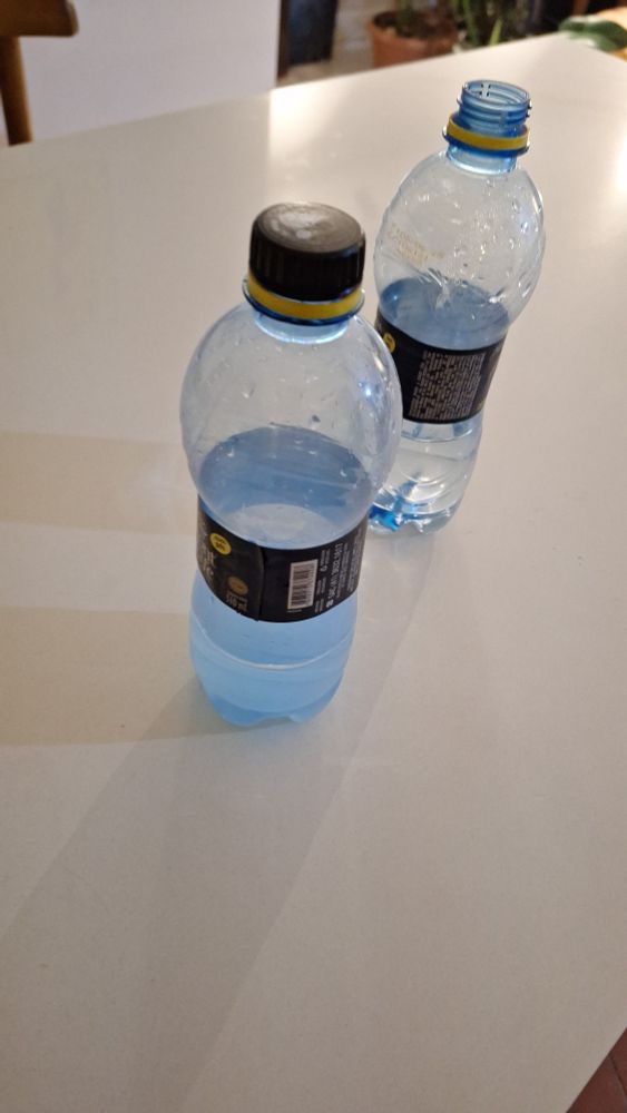 Duas garrafinhas de agia de 500ml lado a lado numa bancada branca. Uma com líquido azulado, fruto da misturinha de álcool e amaciante e a outra com água. As garrafas são iguais. 