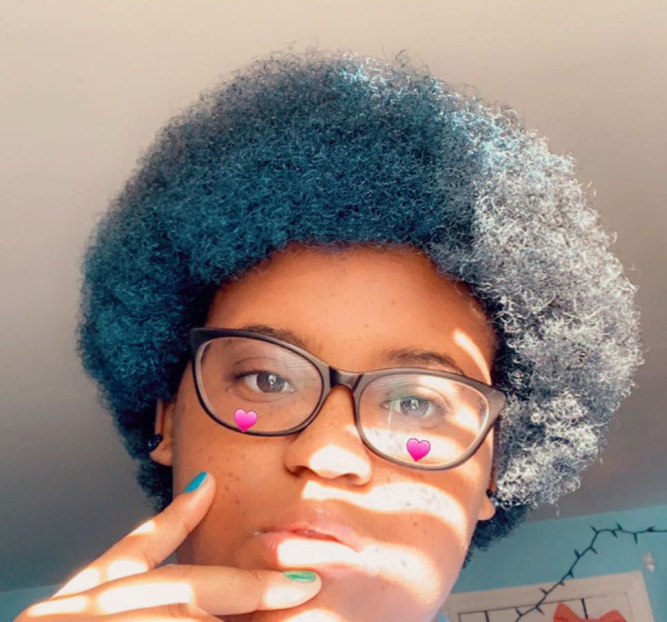 An image of OP with a blue fro and black glasses standing in the sunlight in a blue room.