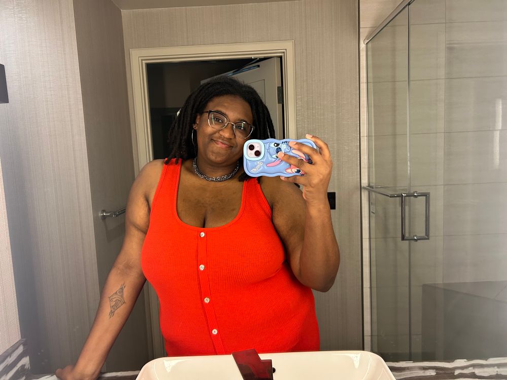 OP in an orange sundress standing in front of a hotel bathroom mirror with a blue stitch cased phone taking the picture. Her locs are much longer in this picture and passed her shoulders.