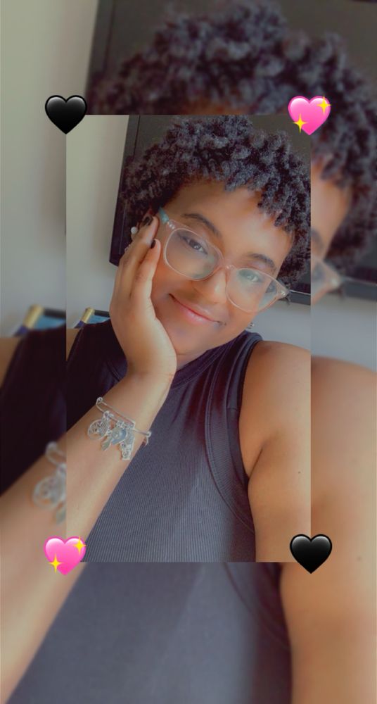 OP with her hand on her chin in a black tank top as she takes a selfie. She has clear glasses. Her locs are still in their twist stage here and are much shorter than the next image. 