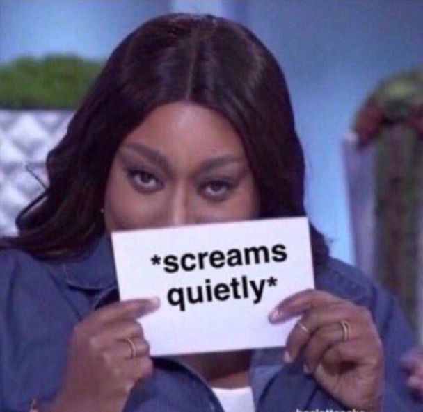 A woman hiding behind a card that says “screams quietly” as she makes a sly face gesturing to the camera