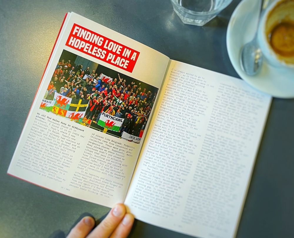 An open copy of Alternative Wales on a table top. The magazine is open on a two-page spread titled ‘Finding Love in a Hopeless Place’ though the article copy is blurred out. There is a large image on the spread of soggy Wales fans in the open away end in Zenica in Bosnia Herzegovina in 2015.
