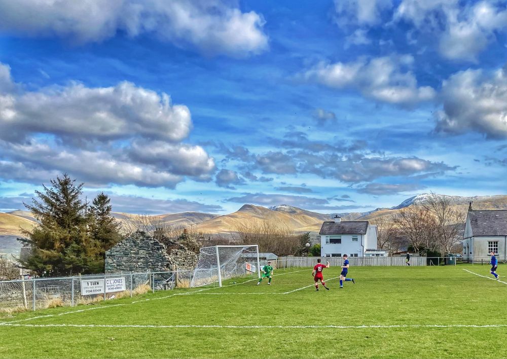 A footballer in maroon volleys a shot over the crossbar from eight yards as a goalkeeper and two defenders in blue look on; beyond the pitch are a white house, a ruined stone outbuilding and the mountainous landscape of Eryri with snow on top of its highest peaks