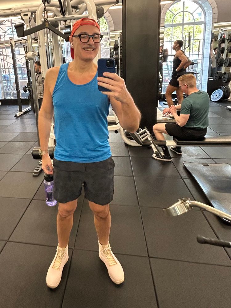 A man (me) is taking a selfie in a gym wearing a blue tank top, black shorts, and white sneakers. He has glasses and a red cap. In the background, other gym-goers are visible using various equipment. The gym has large windows letting light pouring in.