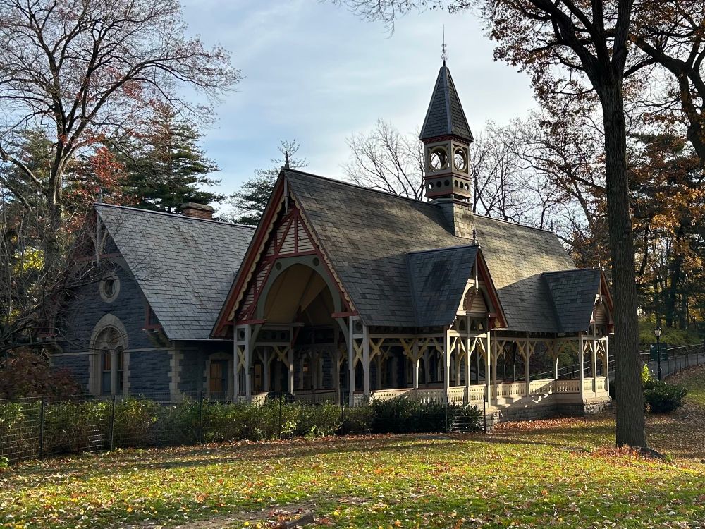 A historic building with Victorian architectural features, surrounded by trees and autumn foliage. It has a steeply pitched roof, decorative trim, and a clock tower. The setting includes a grassy area with scattered fallen leaves.