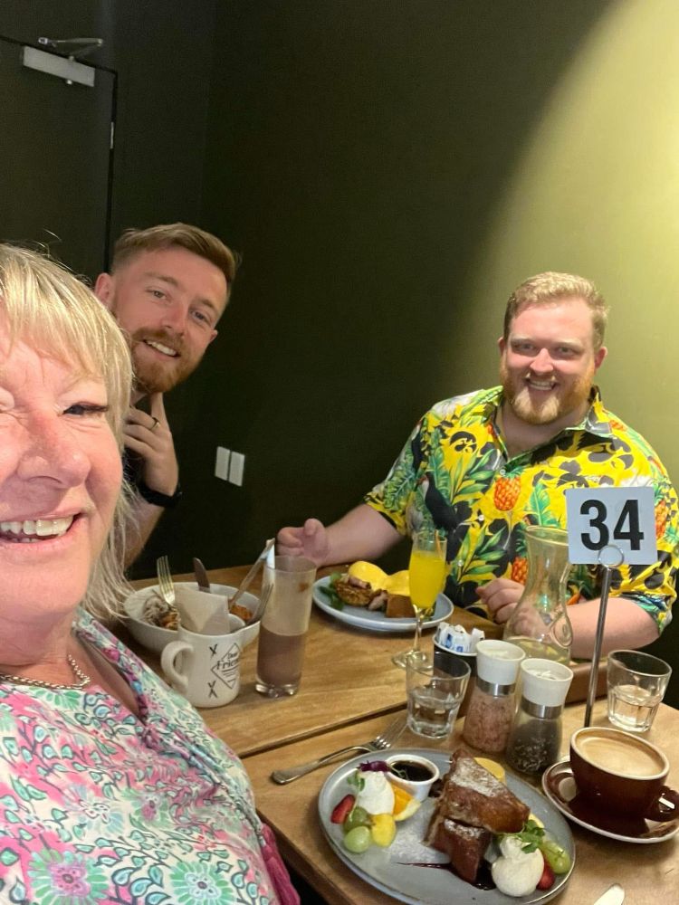 Selfie taken at a Cafe by Shepz’ mum with his brother behind her and Shepz on the other side wearing a yellow summer button up collard shirt covered in a mashup of pineapples, birds & fauna 
