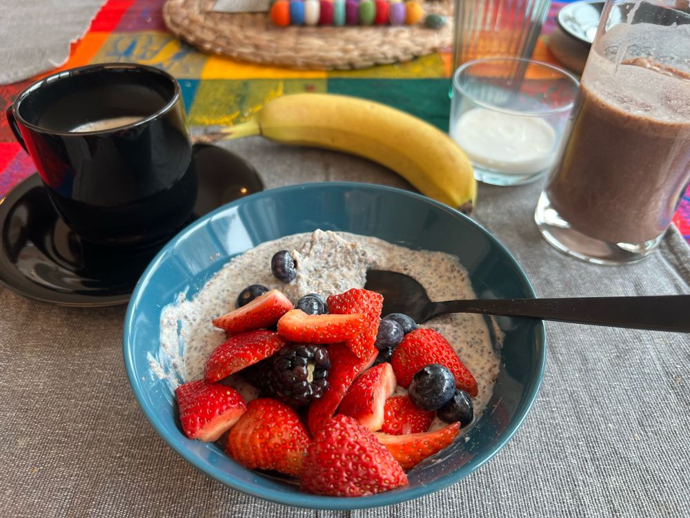 A black cup of coffee, banana, oat creamer, a glass of chocolate protein shake, and a teal bowl of plant-based Greek yogurt with chia seeds and flax seed meal mixed in topped with sliced strawberries, blackberries, and blueberries.