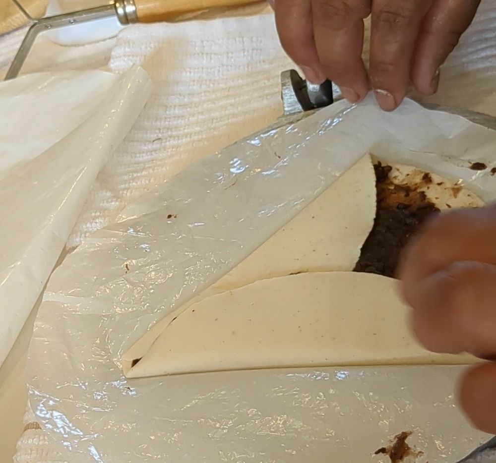 Manos de cocinera plegando una lámina de masa de maíz nixtamalizado para envolver una capa de frijol negro cocinado y apachurrado.