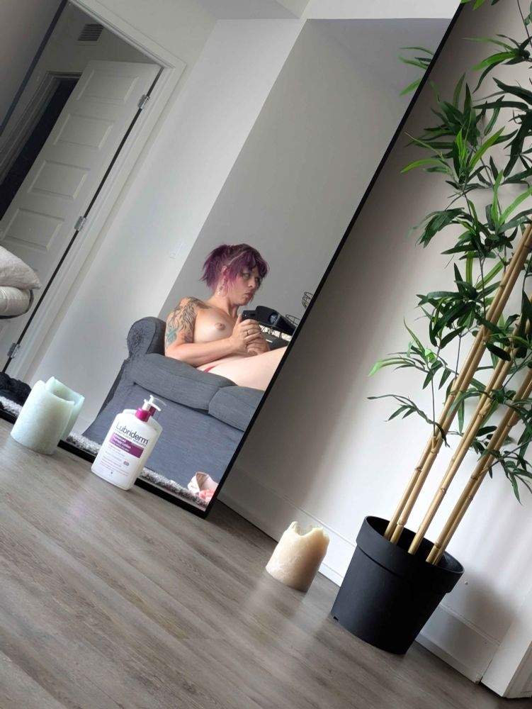 Zoe lounges on a couch, seen through a mirror. She's topless and looking off to one side, her purple hair in a ponytail. 