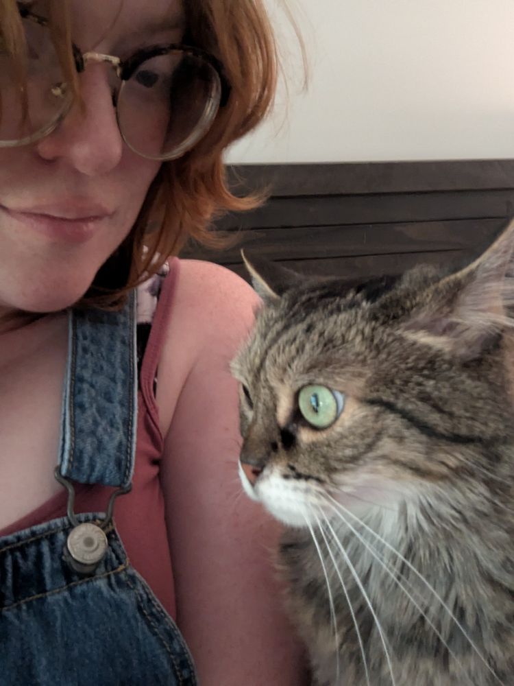 A redheaded person in denim overalls and a dusty rose tank top with bigass glasses sits to the left of a long-haired tabby cat who's clearly up to something.