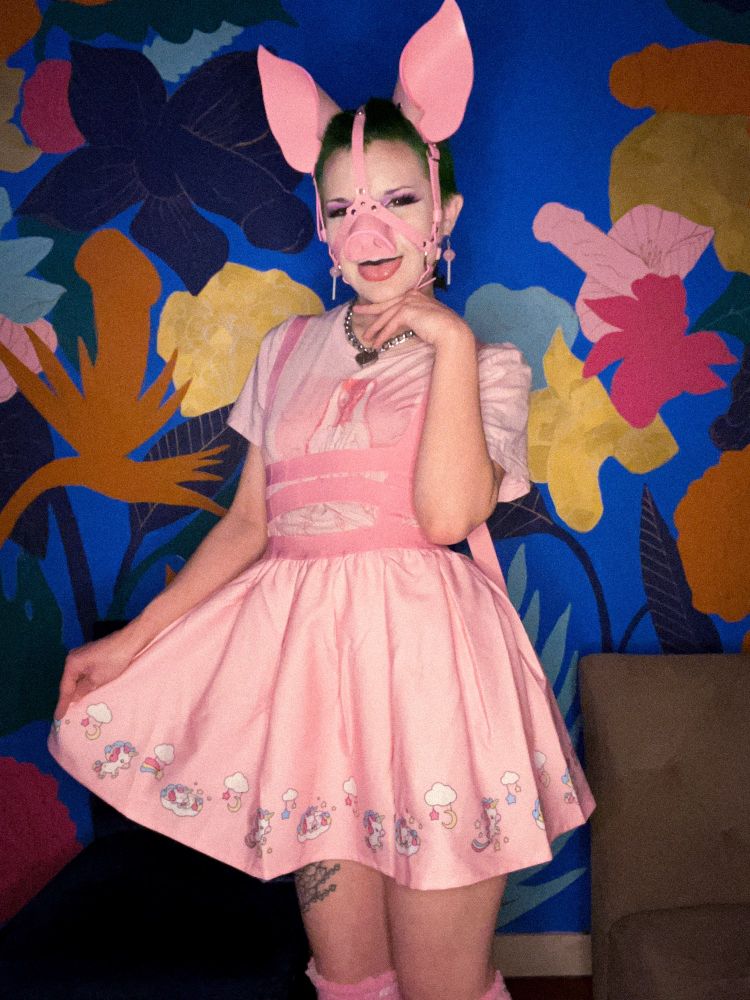 Gwen poses in front of a mural with flowers in the shapes of penises and vulvas. They smile adorably and stick their tongue out while wearing their brand new leather piggy head harness, complete with a snout and big floppy ears. They wear all baby pink with a harness dress, lollipop dangly earrings, and knee-high fuzzy socks. They pull out one end of their dress to show off the bottom which is lined with unicorns and clouds. 