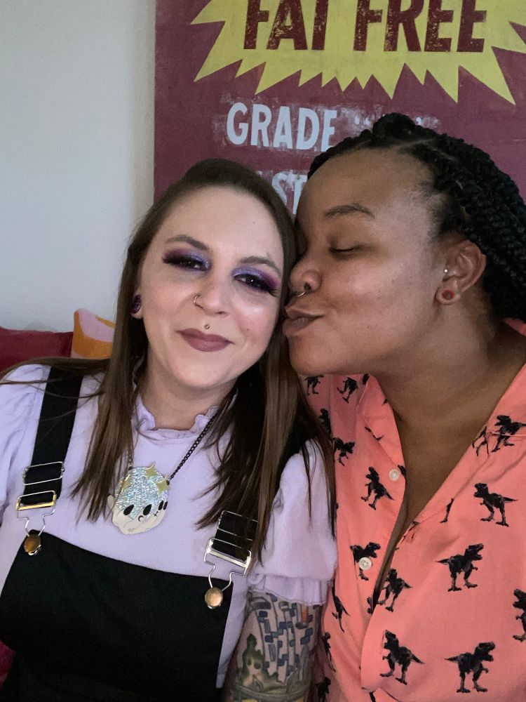 Two adorable femmes, one white and one Black, being super cute and giving a smooch on the cheek. 