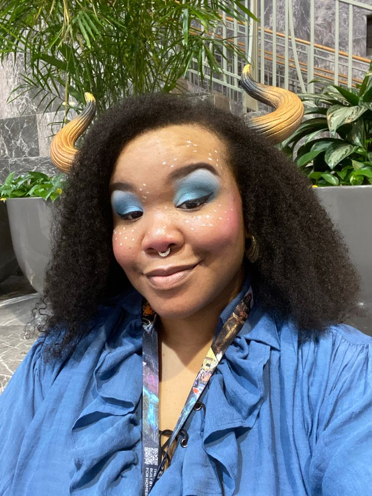 A Black enby at PAXU 2025 dressed as a minotaur with golden horns, blue eyeshadow, kinky hair, and white freckles. 