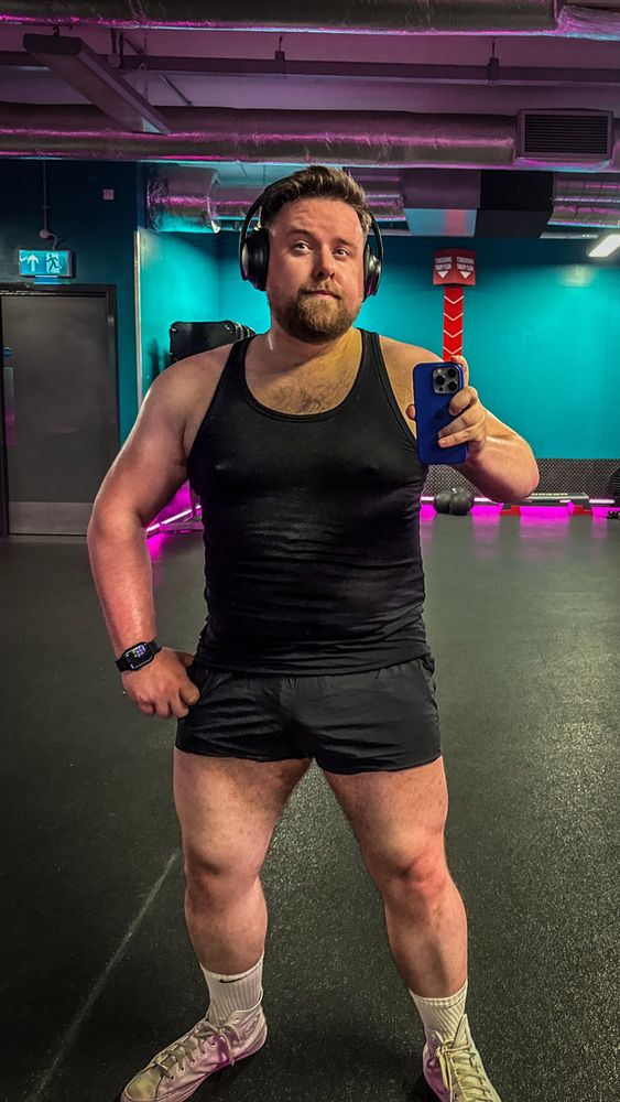 Man in a gym taking a mirror selfie post-workout. He’s wearing a black tank top and short athletic shorts, with visible sweat showing effort. His build is solid and muscular, particularly in the arms and legs. He’s also wearing over-ear headphones, a smartwatch, and light-colored training shoes with white crew socks. The gym background features teal walls, exposed piping, and pink neon lights.