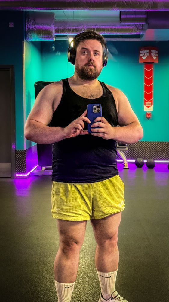 Man taking a gym mirror selfie, wearing a black tank top, yellow athletic shorts, white Nike socks, and sneakers. He has a muscular build with visible arm and leg definition, and a full beard. He is holding a phone and wearing large black headphones, with gym equipment and purple lighting in the background.
