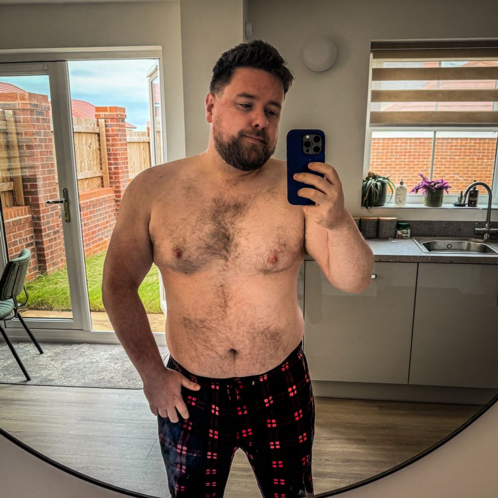 Man taking a mirror selfie in a modern kitchen, shirtless and wearing plaid pajama pants. He has a beard and short hair, with a relaxed expression. His build is broad