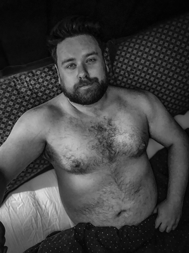 A bearded man with a broad frame and visible chest and abdominal hair lies shirtless in bed, partially covered by a dotted blanket. He has a relaxed, confident expression, with his arm resting at his side. The image is in black and white, adding a moody, intimate tone.