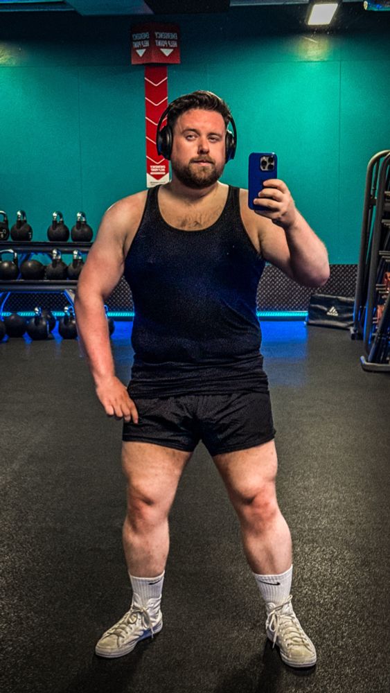 A man takes a gym mirror selfie while wearing a black tank top, short black gym shorts, white Nike socks, and white Converse sneakers. He has a beard, short styled hair, and is wearing black over-ear headphones. His build is visibly muscular, especially in the arms and legs, with a solid and powerful frame. Behind him are rows of kettlebells and a teal-colored wall with emergency signage. The lighting emphasizes his physique and the gym’s sleek, modern vibe. 