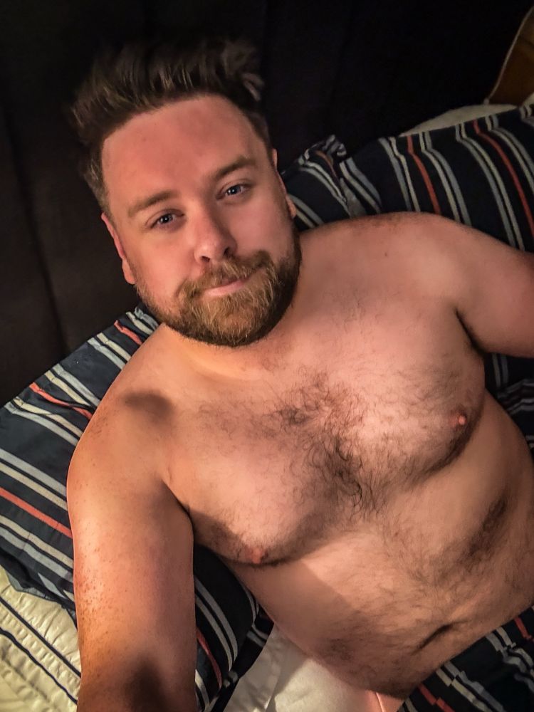 A shirtless man with a trimmed beard and short, styled hair lies on a bed with striped bedding. He has a relaxed facial expression and some chest and body hair. The lighting is warm and soft, and there is no blue sky visible in the image.