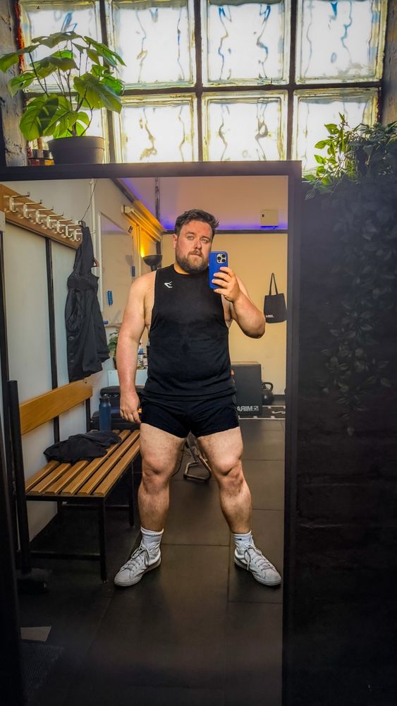 A man stands in a gym locker room taking a mirror selfie. He is wearing a black sleeveless athletic shirt, black shorts, white crew socks, and white sneakers. He has a muscular and solid build, with particularly well-developed thighs and arms. The lighting highlights his muscle definition, especially in the legs and shoulders. Behind him is a wooden bench, gym bags, and a glass-brick window letting in diffused daylight, with some green plants placed above the mirror.