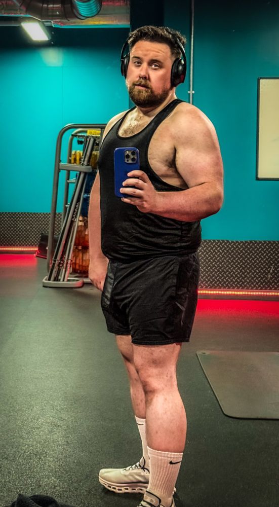 A well-built man with visible muscular development, particularly in his shoulders, arms, and legs, stands in a gym taking a mirror selfie. He is wearing a black tank top that highlights his broad chest and defined upper body, along with black shorts, white Nike socks, and athletic shoes. He has a beard, is wearing over-ear headphones, and has a focused expression. The gym has teal blue walls, equipment in the background, and red accent lighting along the floor.