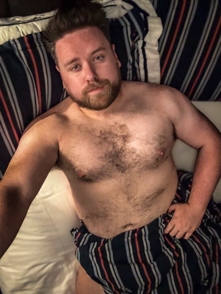 A bearded man with tousled hair is lying shirtless on a bed, partially covered by a striped blanket. He is looking at the camera with a relaxed expression. The bedding features dark blue, white, and red vertical stripes. The lighting is warm and soft, giving the photo an intimate, cozy feel.