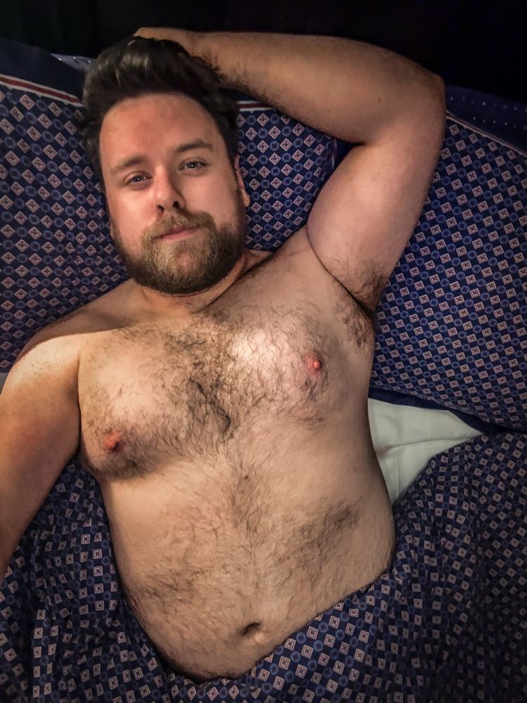 A bearded man with a broad chest and visible body hair lies shirtless in bed, resting one arm behind his head. He has a relaxed expression and is partially covered by a blue patterned blanket and pillow.