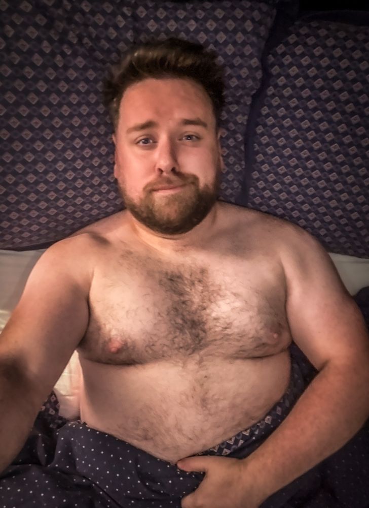 A bearded man with short hair is lying in bed under a dark blue patterned blanket, looking up at the camera with a neutral expression. The bedding and pillows around him have a matching blue geometric pattern. He is shirtless and resting one hand on his stomach.