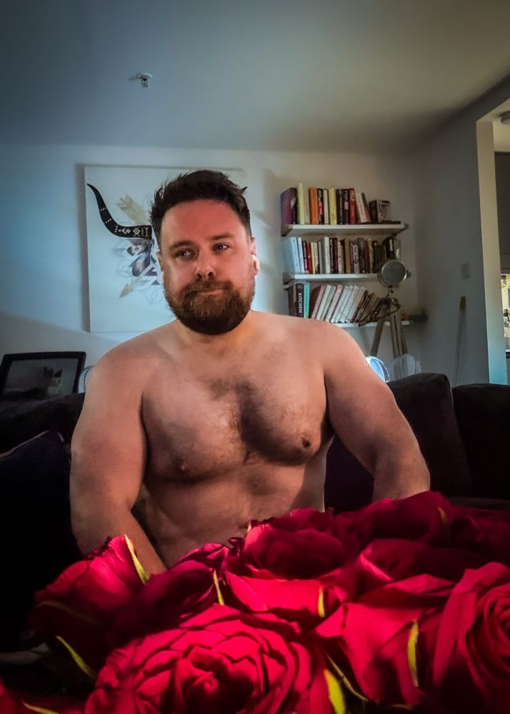 A shirtless man with a muscular and broad upper body, including defined arms and chest, sits on a couch in a softly lit living room. He has a trimmed beard and short hair, and he looks off to the side with a relaxed expression. In the foreground, there is a bouquet of red roses, and behind him are shelves filled with books and wall art featuring a stylized animal skull.