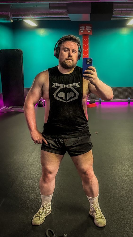 A muscular man stands confidently in a gym, taking a mirror selfie. He wears a sleeveless black tank top with a bold graphic on the chest, short black athletic shorts, and white socks paired with worn light-colored sneakers. His toned arms and strong, defined legs are prominent, suggesting a dedicated weight training regimen. He has a trimmed beard, short hair, and wears large black over-ear headphones. The gym features teal walls, dark flooring, and neon purple lighting along the edges, creating a modern and energetic atmosphere.