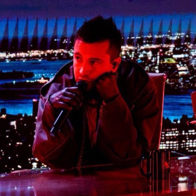 tyler joseph on the tonight show sitting at a desk with his head resting on his hand while singing 