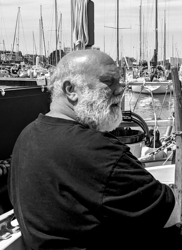 A b&w portrait of an elderly man with a beard, sitting in the cockpit of a small sailboat berthed in a crowded marina..