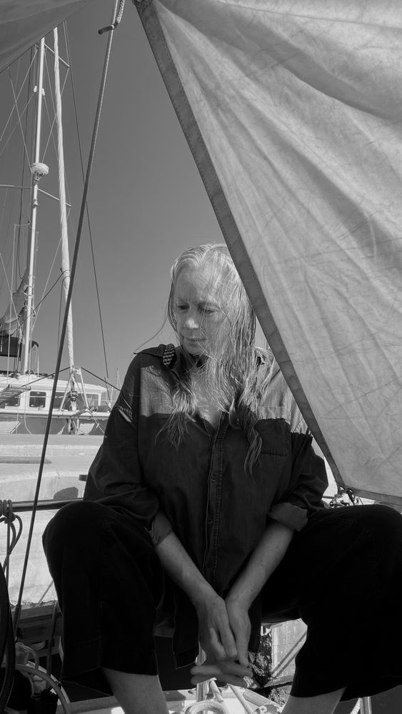 A b&w portriat of an elderly woman sitting under a boom tent on the after-deck of a small sailing boat berthed stern-to a concrete dock.
