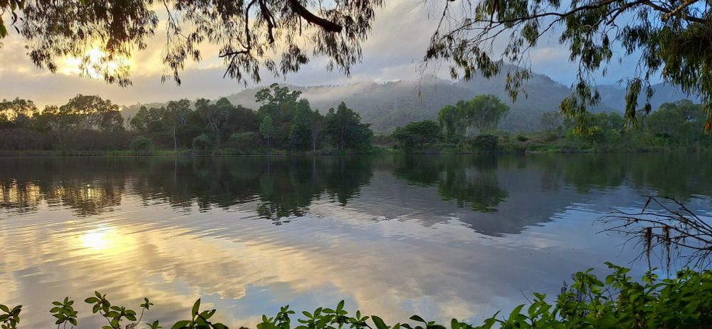 A landscape photo of a river. The sun is about 20° from the horizon on the left hand side. It is obscured by clouds and has an orange hue. There are mountains which have some fog on them spanning from the middle to the right of the image. The river itself takes up majority of the frame and in its reflection you can see the sun and mountains ad well as the trees that line it's bank.