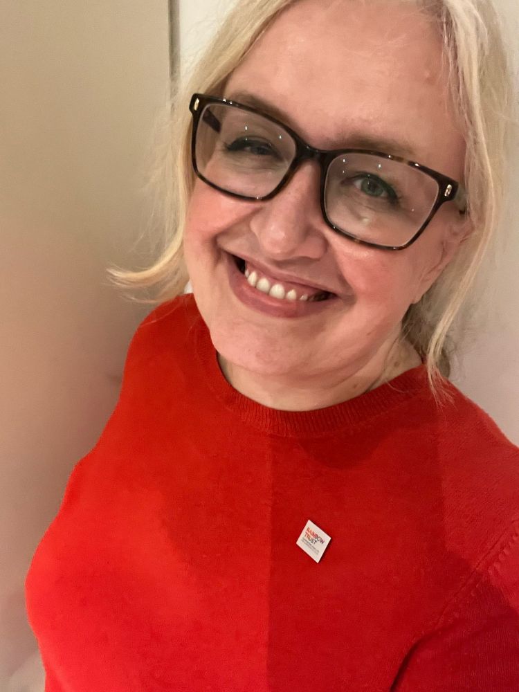 Laura is wearing a red jumper and a Rainbow Trust badge, she is smiling at the camera.