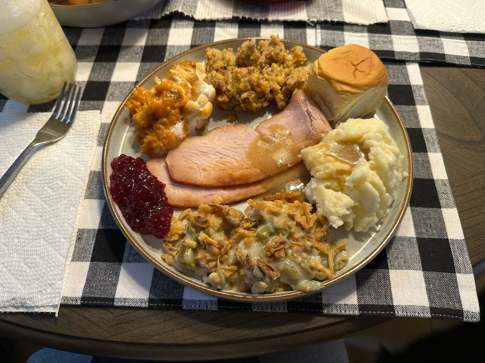 Thanksgiving plate full of turkey, mashed potatoes with some gravy, green bean casserole, cornbread stuffing, sweet potato casserole with little marshmallows, a Hawaiian dinner roll and a small portion of cranberry goo