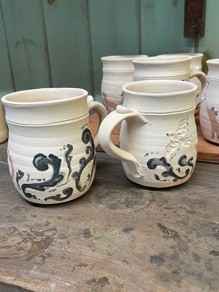 Two unglazed clay mugs. They have dark glaze painted in swirling rosemaled designs. 