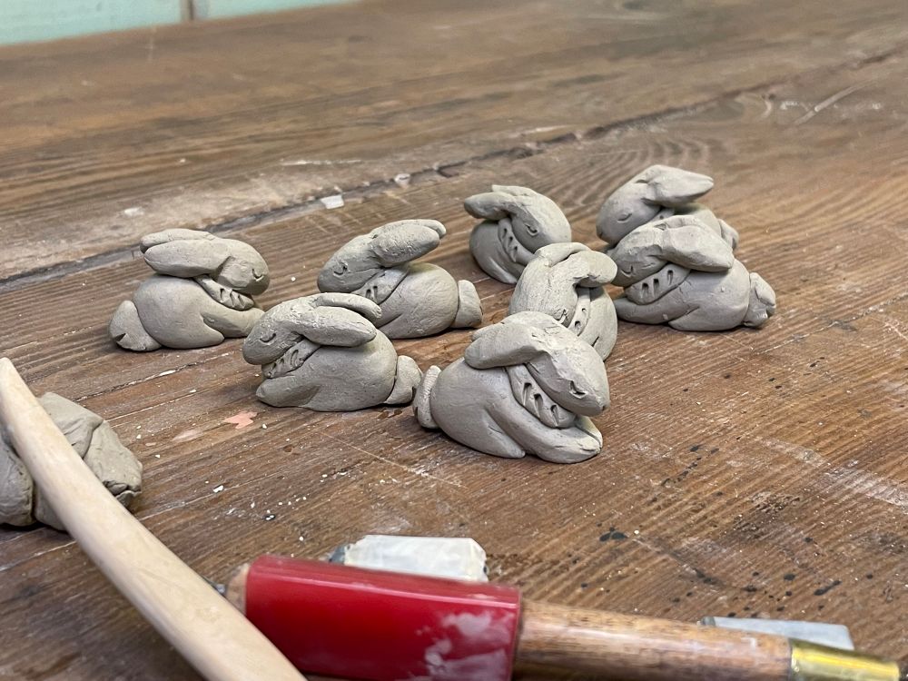 A picture of clay rabbits on a wooden table. The rabbits are small and are made to look as though sleeping. 