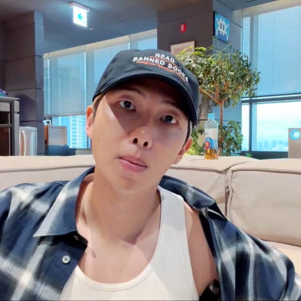 Namjoon during his live today, wearing a white tank top and a blue plaid flannel which is sliding of his shoulder. He has a cap from penguin random house with "Read banned books" written on it.
