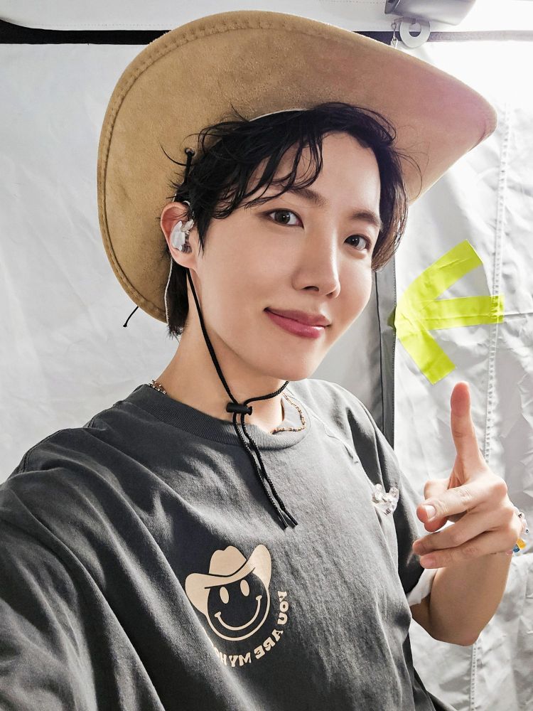Hobi throwing a pesce sign and a half smile while wearing a cowboy hat and a t-shirt with an emoji also wearing a cowboy hat.