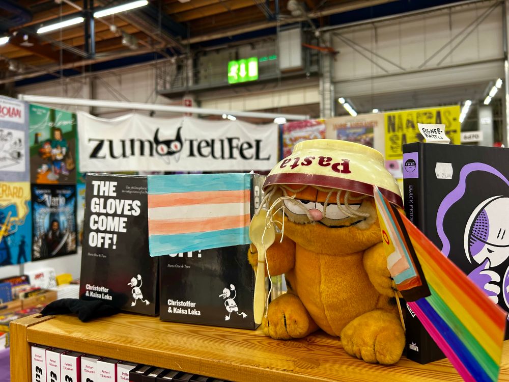 Karvinen-pehmo jolla on pastakulho päähineenä ja joka pitelee trans- ja pride-lippuja. Se on kirjahyllyssä esillä.