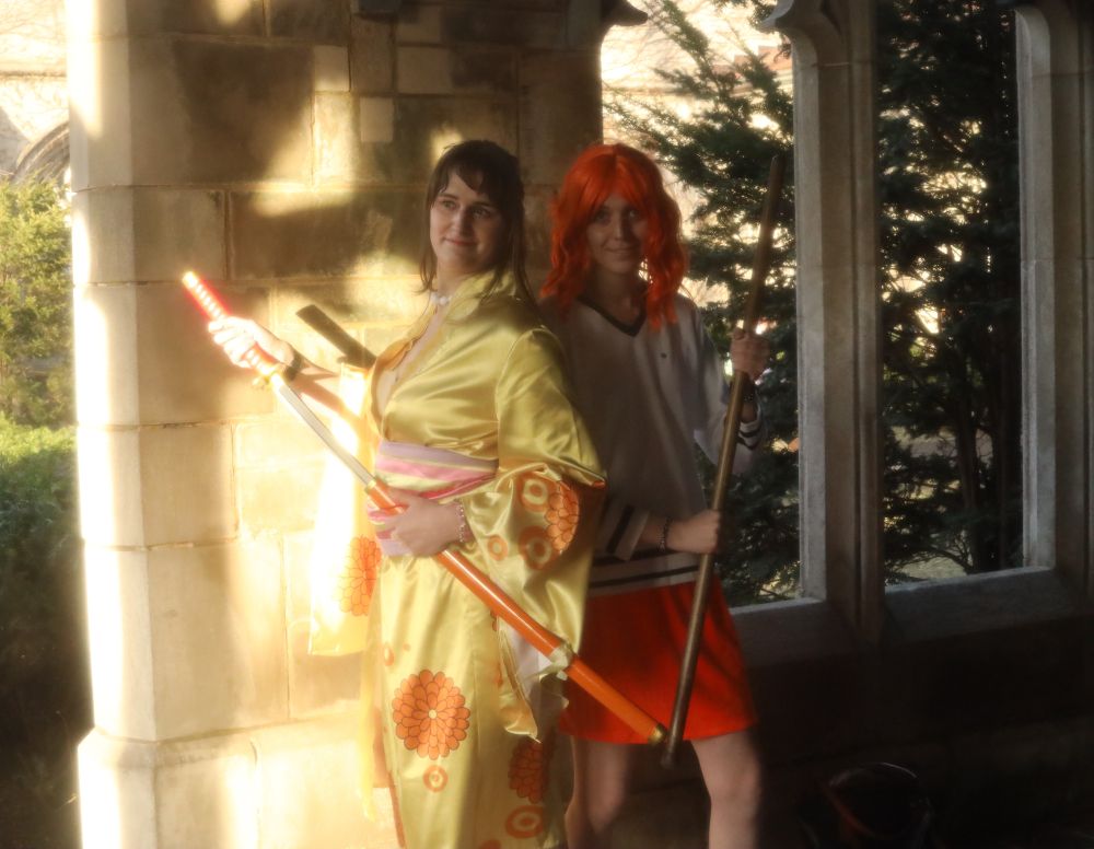 Photo of Norah cosplaying as Kiku from One Piece with Sable cosplaying as Nami from One Piece. Norah is drawing her prop katana while Sable readies her prop staff.