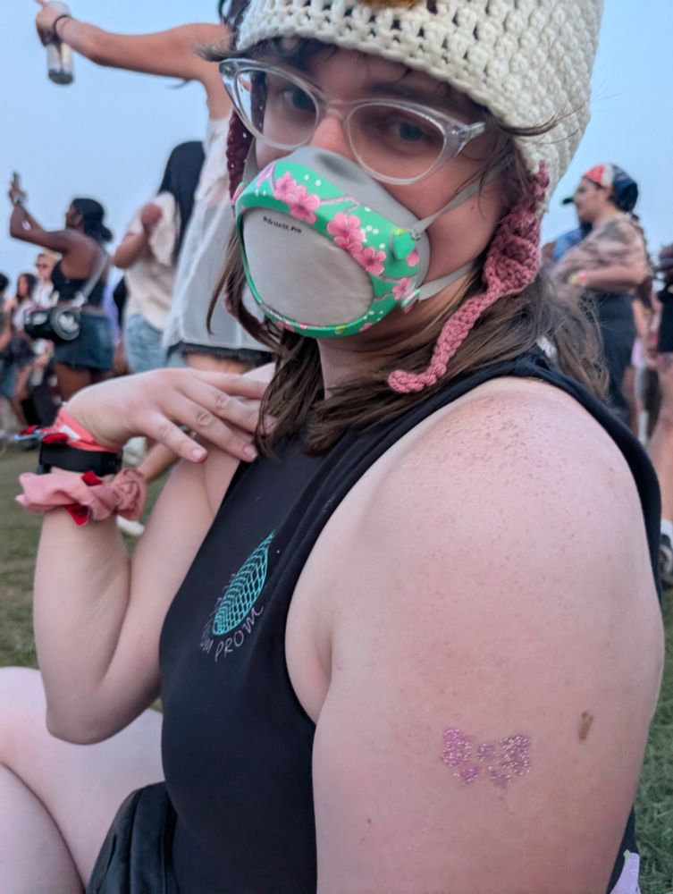 Norah posing with a temporary glitter tattoo of a lavender bow. The tattoo was applied to her during JPEGMAFIA's performance of "i scream this in the mirror before i interact with anyone". She is wearing a Lain Iwakura hat, a cherry blossom Flo Mask and a Beach Bunny T-shirt with the sleeves cut off.