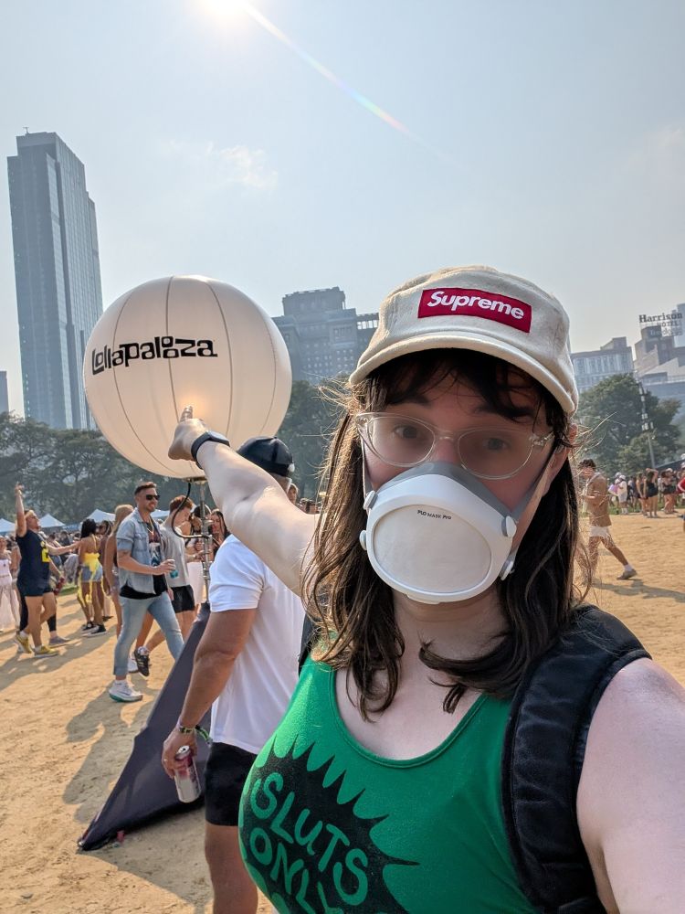 Norah doing a "soyface point" at the Lollapalooza balloon. She is wearing a Supreme hat, a "sluts only" green crop tank, and a flo mask