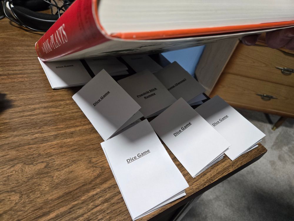 The corner of a wood-panel-topped desk in a room.

Several home-printed booklets with  titles referring to dice and card games are being revealed as sitting underneath a large, broad, hardcover book, which is being lifted up from the bottom edge by an offscreen hand.

The large book is being used to press the booklets flat after folding.