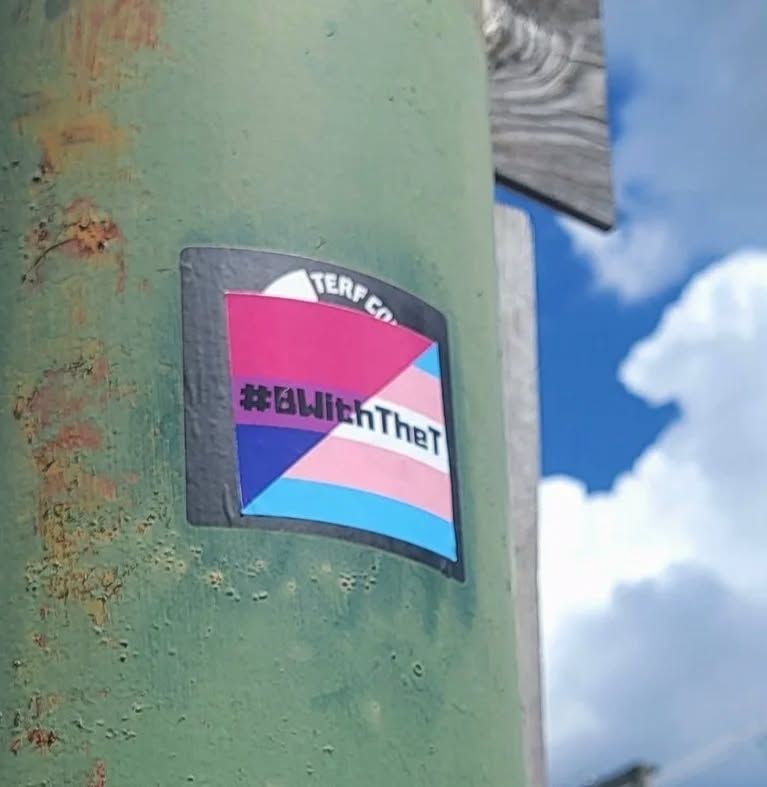 A lamppost with a transphobic sticker on it, which has been covered with a BWithTheT sticker
