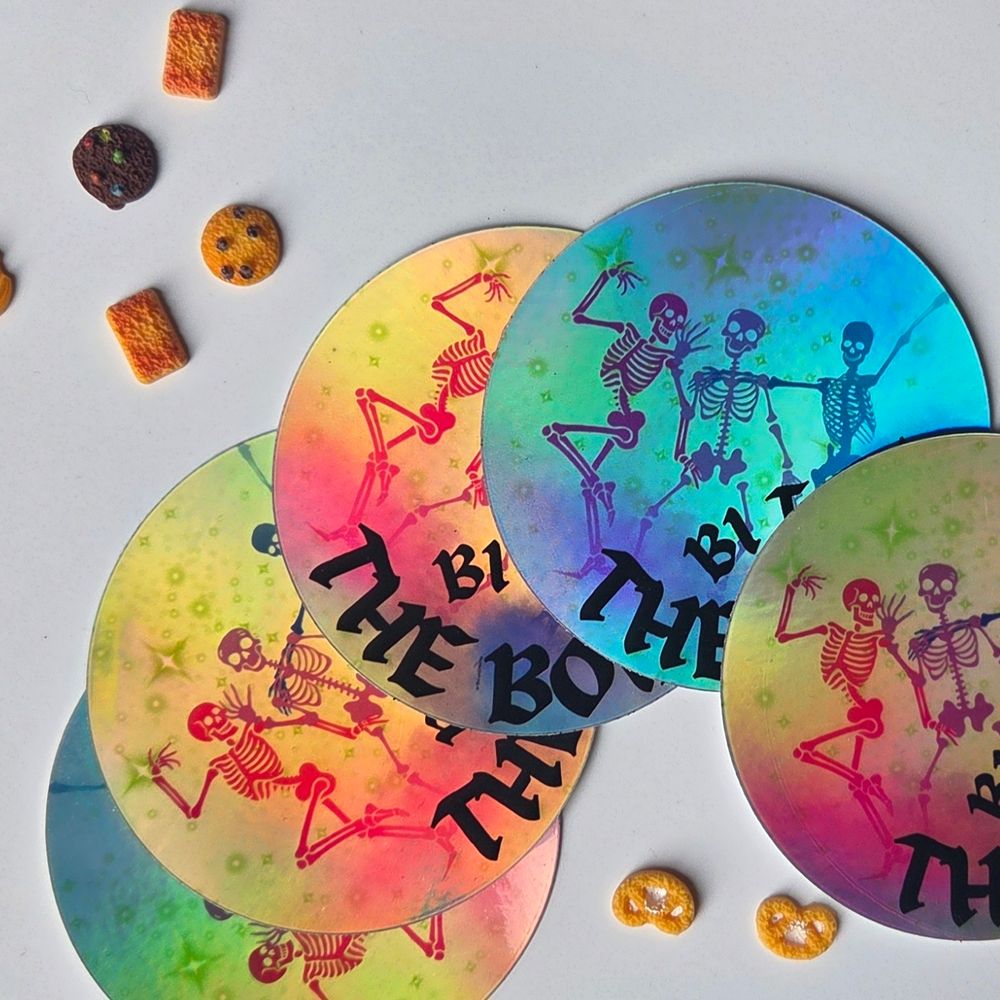 On a white background with Miniature cookies and Biscuits, five iridescent stickers with the image of three dancing skeletons in pink, purple and blue, and the words bi to the bone.