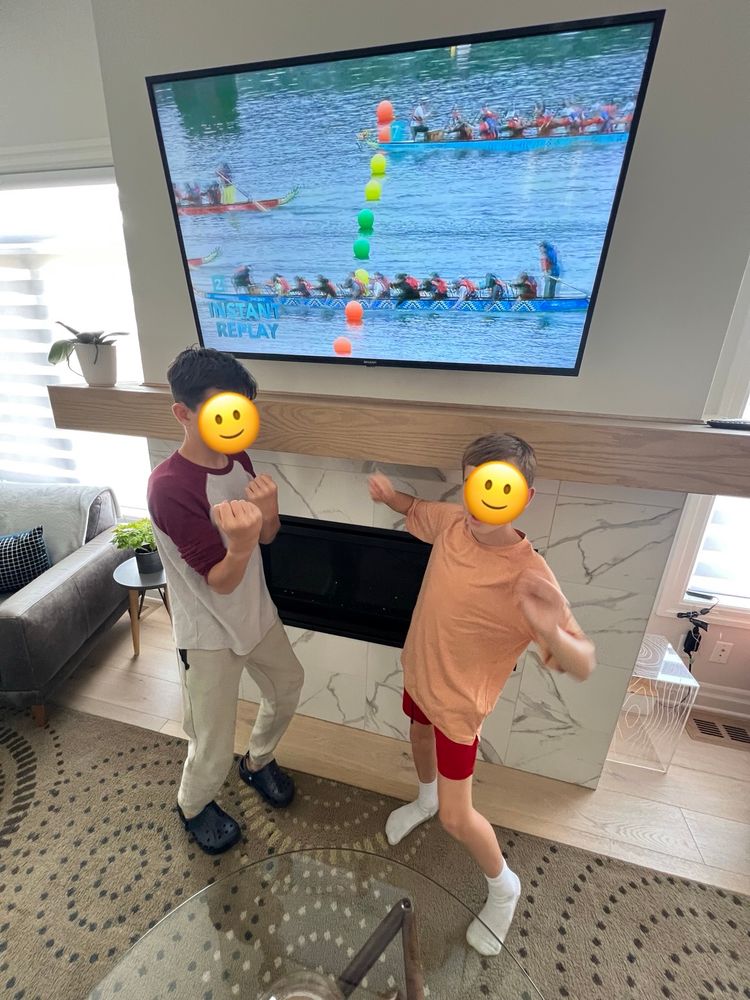Two boys cheer enthusiastically in front of a television showing an instant replay of a dragon boat race. The TV displays multiple dragon boats nearing the finish line, marked by coloured buoys. The boys stand in front of a modern fireplace, visibly excited as they support their mom’s team from home